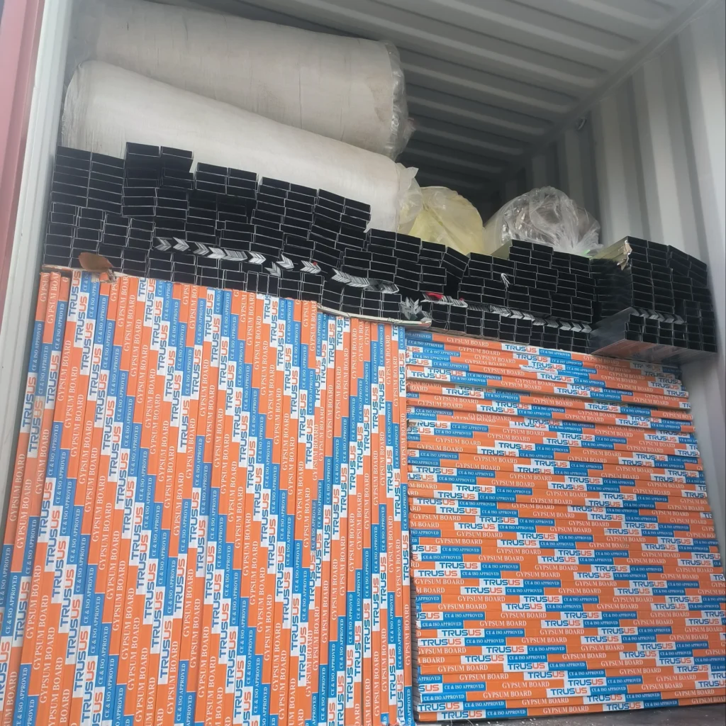 A shipping container filled with stacks of drywall labeled 'Trusus' and black metal frames, with rolls of material on top.