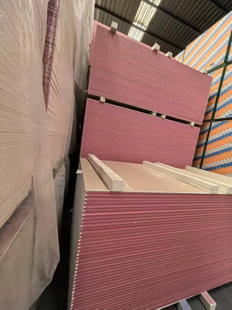 Large stacks of pink drywall sheets neatly arranged in a warehouse. The warehouse has a metal roof and is well lit.