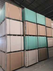 Multiple stacks of drywall panels in various colors, including beige and green, organized in a warehouse with a metal roof.