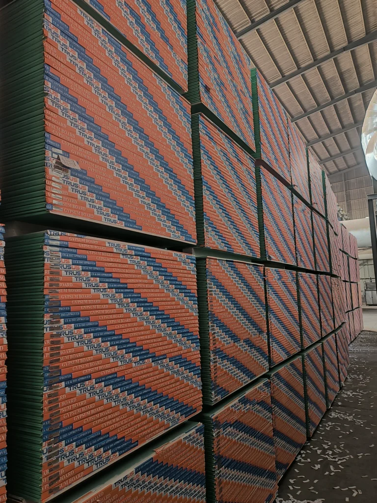 Large stacks of gypboard panels labeled 'TRUSUS' arranged in a warehouse with a high ceiling, showing organized industrial storage.