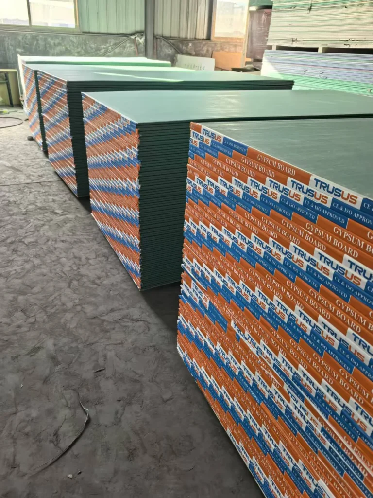 Piles of gypsum boards stacked neatly in a warehouse, with labels visible on the side and ample natural light from windows.