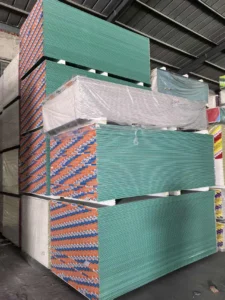 Large stacks of drywall sheets, wrapped and stored in a warehouse setting, with visible green and white panels and colorful branding.