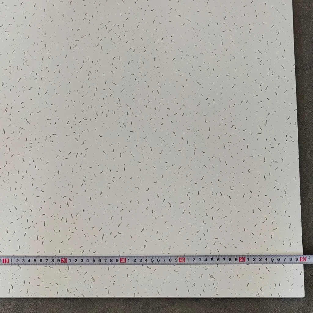 Acoustic ceiling tile featuring small indents and patterns, measured with a tape measure along one edge on a dark surface.