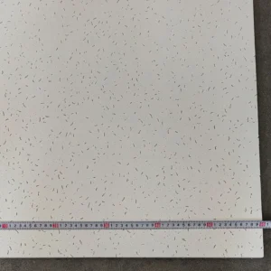 Acoustic ceiling tile featuring small indents and patterns, measured with a tape measure along one edge on a dark surface.
