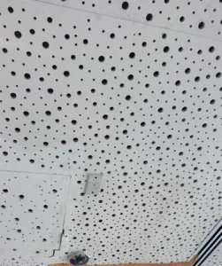 A perforated acoustic ceiling panel with visible holes, a mounted control device, and a section covered with plastic for repairs.